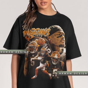 May include: Black t-shirt featuring a collage of a football player in action, with the name "Higgins" in an orange, distressed font. The design includes multiple images of the player in his uniform, with the number 5.