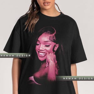 Puede incluir: Camiseta negra con un retrato rosa de una persona sonriente. El retrato es un primer plano de la cara y la parte superior del cuerpo de la persona. El texto "NAWAW DESIGN" está impreso en la camiseta.