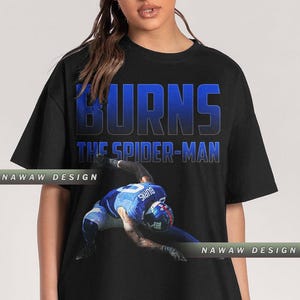 May include: Black t-shirt featuring the text "BURNS THE SPIDER-MAN" in blue. The shirt displays an image of a football player in a blue uniform. The design includes the name "BURNS" on the player's jersey.