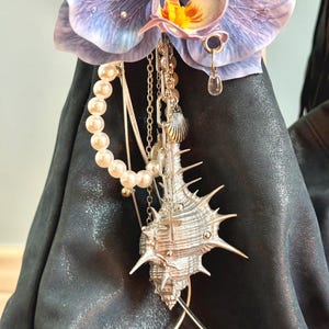 May include: A decorative orchid flower with purple petals and a yellow centre. A silver seashell pendant with a cross hangs from the flower, along with a pearl strand and various chains. The background is a dark, textured surface.