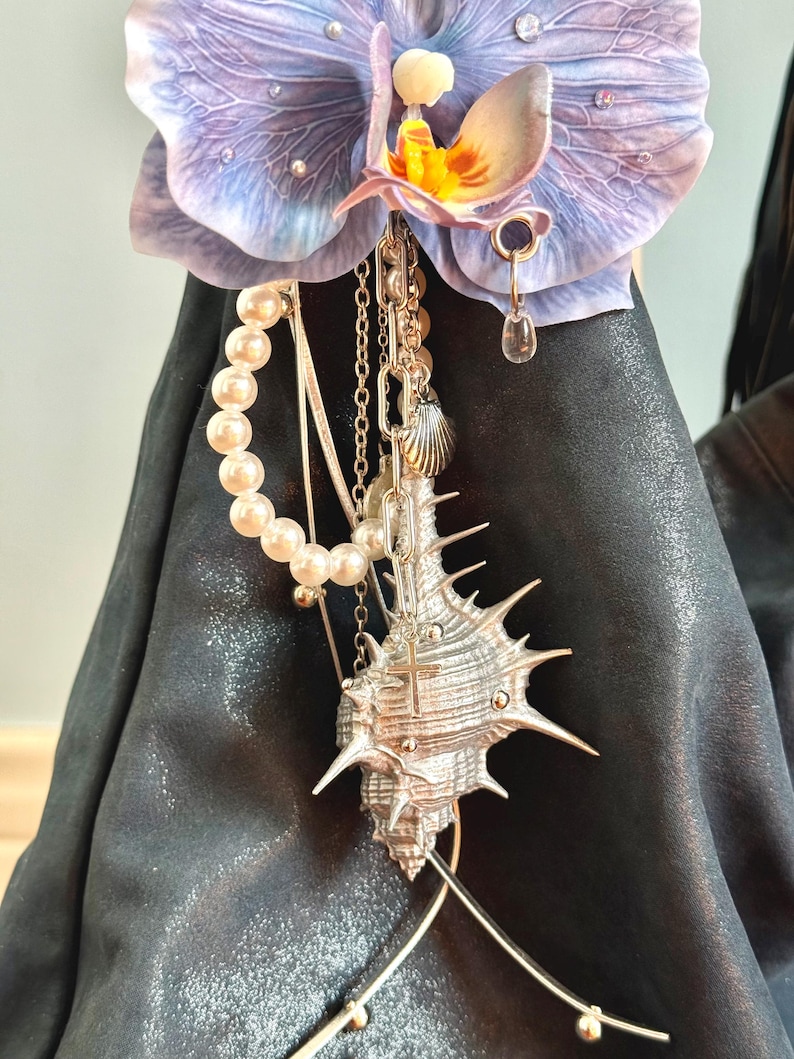 May include: A decorative orchid flower with purple petals and a yellow centre. A silver seashell pendant with a cross hangs from the flower, along with a pearl strand and various chains. The background is a dark, textured surface.