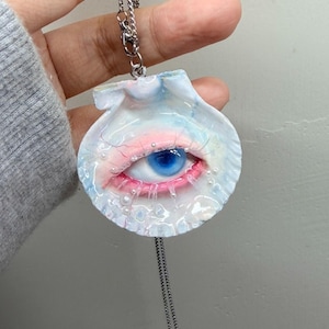 Op de afbeelding: Een unieke hangerketting met een schelpmotief met een blauw oog en roze accenten. Het oog is gedetailleerd met een blauwe iris en rode wimpers. De hanger hangt aan een zilveren ketting.