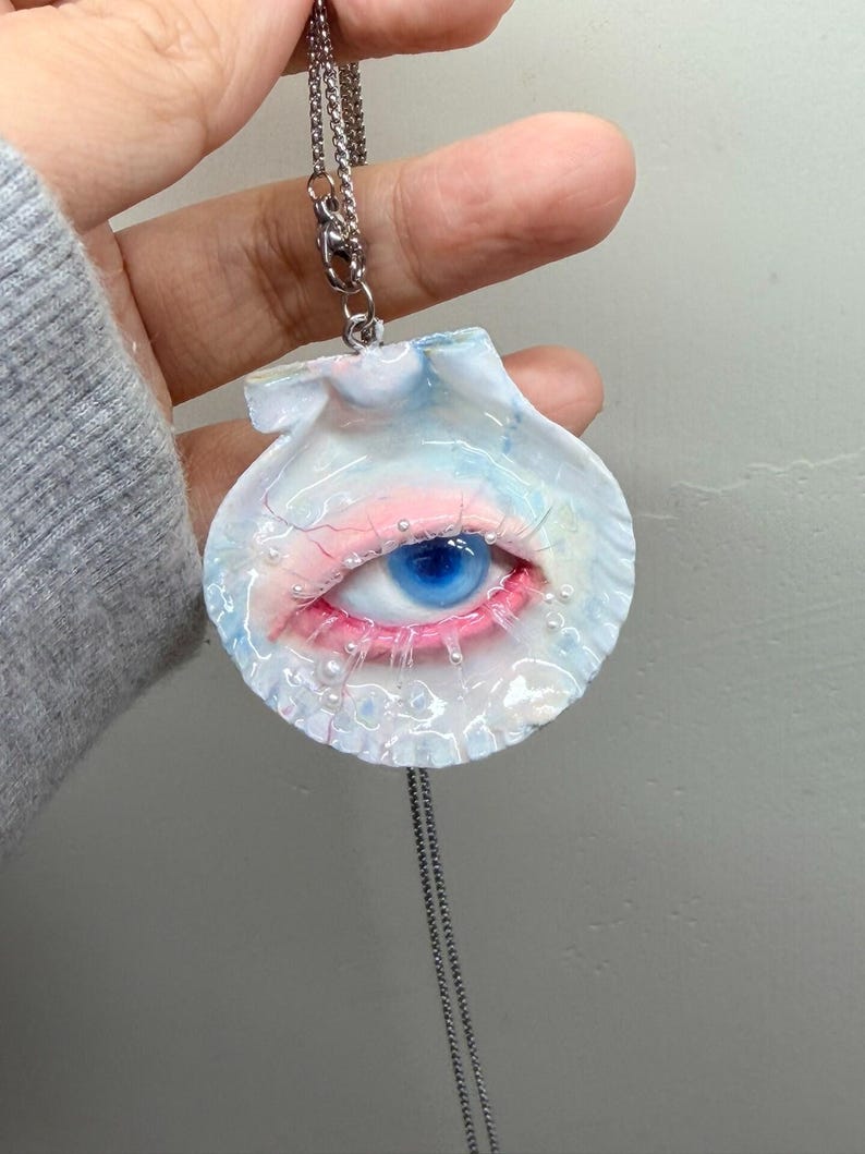 May include: A unique pendant necklace featuring a seashell design with a blue eye and pink accents. The eye is detailed with a blue iris and red eyelashes. The pendant hangs from a silver chain.