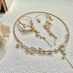May include: A gold necklace and earrings set with pearl accents. The necklace is a delicate hoop with a dangling pearl chain. The earrings are a single hoop with a dangling pearl chain.