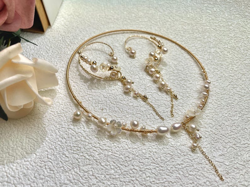 May include: A gold necklace and earrings set with pearl accents. The necklace is a delicate hoop with a dangling pearl chain. The earrings are a single hoop with a dangling pearl chain.