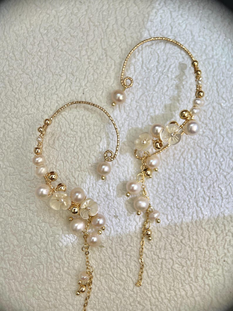 May include: A pair of gold ear cuffs with pearl and flower accents. The cuffs are made of thin wire and have a delicate, feminine look.