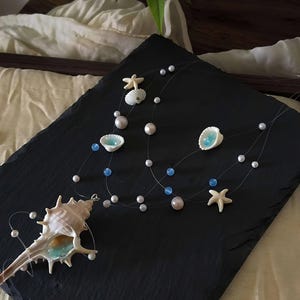 May include: A delicate necklace featuring seashells, starfish, and pearls. The necklace has a transparent string with white and blue beads. The seashells have a light blue interior. The necklace is displayed on a black surface.
