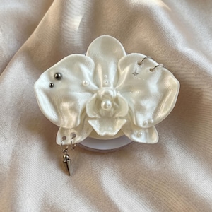 Könnte beinhalten: Ein weißer, orchideenförmiger Handygriff mit Perlglanz-Finish, verziert mit kleinen eingebetteten weißen Perlen, silberfarbenen Metallringen, zwei dunklen kugelförmigen Nieten, einem kleinen vierzackigen Sternanhänger und einem hängenden Metall-Spike.