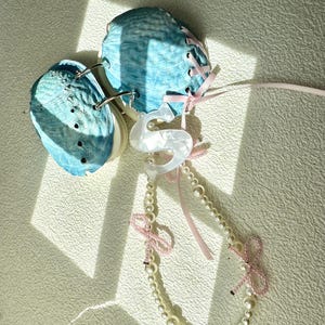 Puede incluir: Un diario hecho a mano con cubiertas de conchas marinas azules, atado con cinta rosa. Un collar de perlas con un colgante de cisne blanco y lazos rosas está adjunto. El diario está abierto, revelando las páginas interiores.