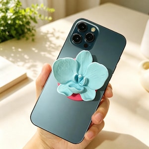 May include: A dark green smartphone featuring a three-lens camera system on the back. Attached to the centre of the phone is a circular phone grip with a three-dimensional, light blue orchid flower design mounted on a solid pink circular base.