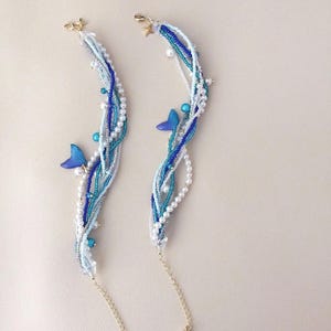 May include: Two beaded bracelets with a mermaid theme. Each bracelet features strands of white, light blue, and dark blue beads, accented with small blue butterfly charms and gold-colored star clasps. The bracelets have a delicate, elegant design.