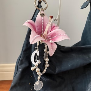 Puede incluir: Un dije en forma de lirio rosa con detalles de perlas y un collar de perlas cuelgan de un bolso azul oscuro. El dije está sujeto a un anillo plateado. El bolso tiene una superficie texturizada y está adornado con pequeños tachuelas doradas.