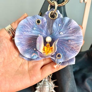May include: A detailed, blue and purple orchid flower charm with a pearl centre and clear crystal accents. The charm is attached to a gold-tone ring and a silver seashell charm. The orchid is approximately 10 cm wide.