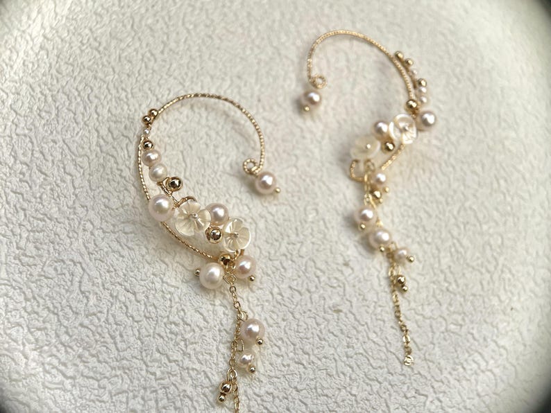 May include: A pair of gold ear cuffs with pearl and white flower accents. The ear cuffs are designed to wrap around the ear and have a dangling chain with pearls.
