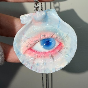 May include: A unique pendant necklace featuring a seashell design with a painted eye. The eye has a blue iris, pink and red accents, and is surrounded by small, pearl-like embellishments. The pendant hangs from a silver chain.