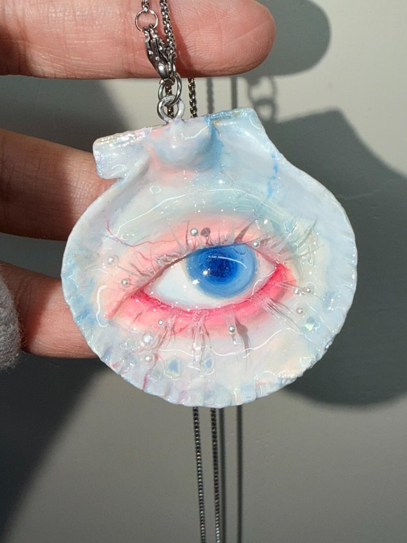 May include: A unique pendant necklace featuring a seashell design with a painted eye. The eye has a blue iris, pink and red accents, and is surrounded by small, pearl-like embellishments. The pendant hangs from a silver chain.