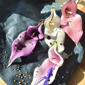Pode incluir: Arranjo de lírios-calla estilizados em tons de roxo, branco e rosa, com caules verdes. As flores são adornadas com ilhós e tachas metálicas, e uma fita roxa. O fundo é um material azul escuro semelhante a couro.