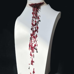 May include: A choker necklace featuring a sheer band with a dense, crescent-shaped cluster of dark red and black beads. A long, cascading fringe of delicate threads hangs from the center, adorned with various red and black beads and crystals.