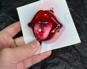 May include: A red, glossy, sculpted mouth with a tongue sticking out and a silver stud piercing. The tongue is pink and white with a black line down the center.