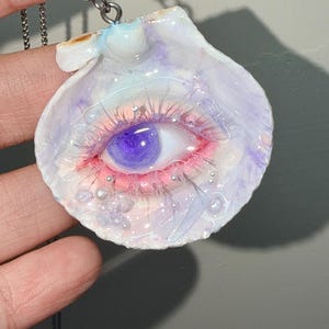 May include: A unique pendant necklace featuring a seashell design with a detailed eye. The eye has a purple iris, pink and red accents, and delicate eyelashes. The seashell is white and lavender, with silver-colored embellishments.