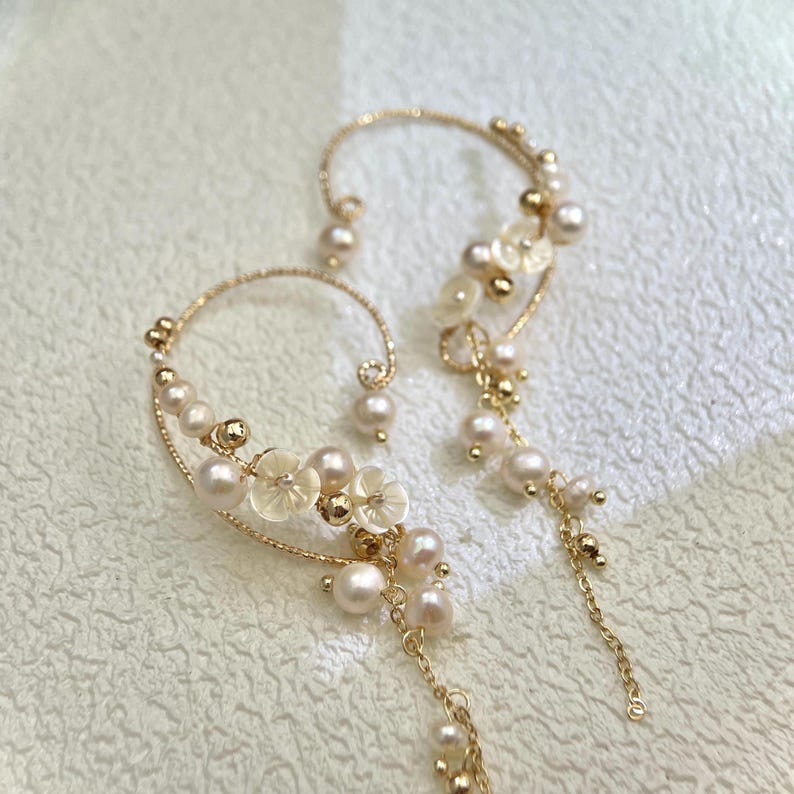 May include: A pair of gold ear cuffs with delicate pearl and flower accents. The cuffs are designed to wrap around the ear and feature a dangling chain with additional pearls.