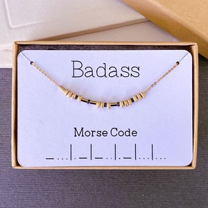 May include: A white card with the word "Badass" printed in black. A gold chain necklace with black beads is displayed on the card. The card also includes the text "Morse Code" and the code for the word "Badass" in Morse code.