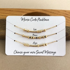 以下が含まれることがあります： 白いカードに「Morse Code Necklace」と「Choose your own Secret Message」と書かれた、黒いビーズで「Love」、「Peace」、「Hope」の文字を綴った3本の金色のネックレス。