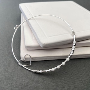 May include: A silver bangle bracelet with a heart charm and small silver beads. The beads have a textured surface.