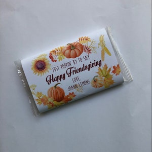 May include: A wrapped candy bar with a Thanksgiving theme. The wrapper features pumpkins, sunflowers, and fall leaves. Text on the wrapper reads "Just Poppin' By To Say Happy Friendsgiving Love, Joann Lemons."
