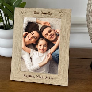 May include: A light wood photo frame with a white mount and a photo of a family of three. The frame has the words "Our Family" engraved on the top and "Stephen, Nikki & Ivy" engraved on the bottom.