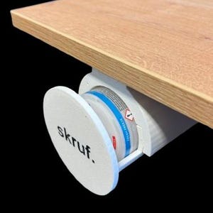 Puede incluir: Organizador de escritorio blanco con una tapa redonda blanca que dice "skruf". El organizador está unido a la parte inferior de un escritorio de madera.