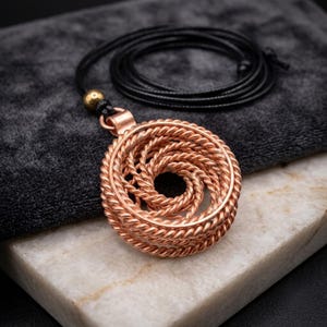 May include: A circular pendant crafted from copper-colored metal wire twisted into a dense, spiraling coil pattern. The pendant is attached to a thin, black cord necklace that features a small, round, metallic bead near the top attachment point.