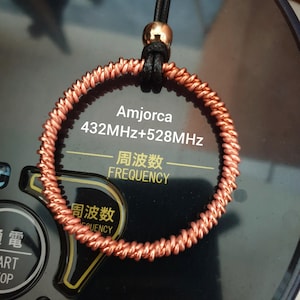 Pode incluir: Um colar com um pingente de fio de cobre em forma de círculo, um cordão preto e uma conta dourada. O pingente tem o texto "Amjorca 432MHz+528MHz 频率 FREQUENCY".