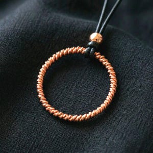 Peut inclure: Un pendentif circulaire fabriqué en fil de cuivre torsadé, suspendu à un cordon noir. Le pendentif présente une finition en cuivre métallique et est fixé au cordon par une petite perle ronde couleur cuivre et un nœud noir.
