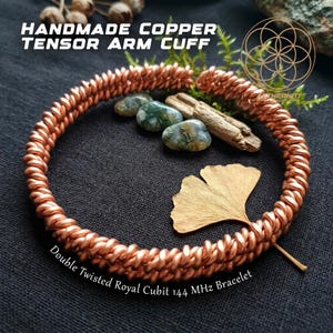 May include: A handmade copper tensor arm cuff, identified as a double twisted royal cubit 144 MHz bracelet. The metallic copper band features a continuous twisted wire design, accompanied by green moss agate stones and a gold-toned ginkgo leaf.
