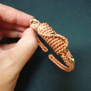May include: A copper wire cuff bracelet featuring a woven, braided texture throughout the band. The design includes three small, smooth, metallic gold-toned spherical beads integrated into the wire structure at intervals along the bracelet.