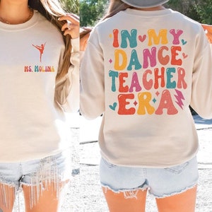 May include: A cream-colored sweatshirt with a ballet dancer graphic and the text "MS. MOLINA" on the front. The back of the sweatshirt features the text "IN MY DANCE TEACHER ERA" in colorful, retro-style lettering.