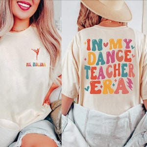 May include: Cream-colored t-shirt with a ballet dancer graphic and the text "Ms. Molina" in rainbow colors. The back of the shirt features the text "In My Dance Teacher Era" in a retro, colorful design. A casual, comfortable style.