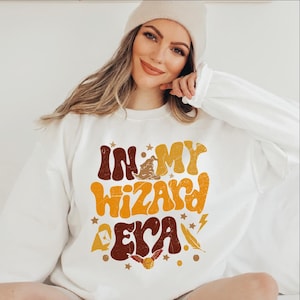 May include: White sweatshirt with a graphic print that says "In My Wizard Era" in a retro font. The graphic is in shades of brown, yellow, and orange and features a wizard hat, a lightning bolt, and a feather.