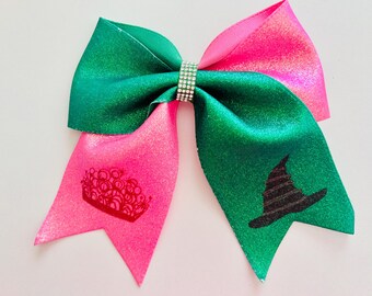 May include: A large, decorative hair bow with a green and pink color scheme. The bow features a rhinestone center and is adorned with a red crown and a black witch hat design. The bow is made of a shiny, glittery material.