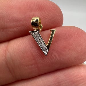 Pode incluir: Um pingente dourado em forma da letra "V". A argola superior e o lado direito do "V" são dourados. O lado esquerdo do "V" é prateado com pequenos detalhes brilhantes. O pingente é segurado entre os dedos.