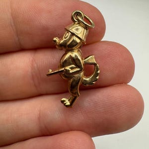 Könnte beinhalten: Ein goldfarbener Anhänger in Form eines Zeichentrickhundes mit Hut und Gewehr. Der Hund hat einen Schwanz und steht aufrecht. Der Anhänger hat oben eine Schlaufe zum Befestigen an einem Armband oder einer Halskette.