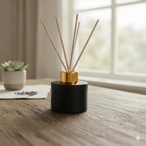 Könnte beinhalten: Ein schwarzer Diffusor für ätherische Öle mit einem goldfarbenen Kragen und Holzstäbchen. Der Diffusor steht auf einem Holztisch, im Hintergrund eine kleine Sukkulente in einem weiß-beigen Topf. Im Hintergrund ist ein Fenster zu sehen.