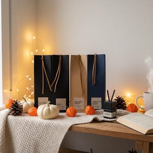 Könnte beinhalten: Herbstliche Dekoration mit drei rechteckigen Schachteln in Schwarz, Beige und Marineblau, jeweils mit Ledergriff. Weiße Kürbisse, orangefarbene Lampions und Tannenzapfen sind auf einem Holztisch mit einer Strickdecke arrangiert. Lichterketten sorgen für einen warmen Schein.