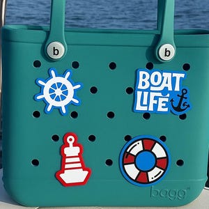 以下が含まれることがあります： 航海をテーマにしたデカールが付いた、ティール色のトートバッグ。バッグにはハンドルがあり、「BOAT LIFE」の文字とアンカーが付いています。その他のデカールには、船の舵、ブイ、救命浮環が含まれます。バッグはボートにあります。