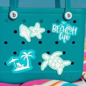 May include: Teal tote bag with white beach-themed stickers, including a seashell, starfish, and turtle. The bag has two handles and the words "BEACH life". The bag is on a striped towel at the beach.
