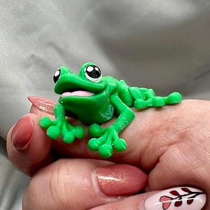 May include: A green frog figurine with large eyes and a wide smile. The frog is sitting on a person's hand.