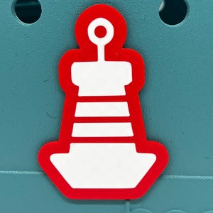 以下が含まれることがあります： 赤と白の灯台型のチャーム。チャームは赤いアウトラインと白い内部を持ち、水平ストライプと円形のトップがあります。チャームは、2つの円形の穴があるティール色の背景に配置されています。