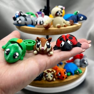 May include: A collection of small, colorful, 3D printed animal figurines, including a ladybug, a squirrel, a chameleon, a turtle, a sloth, a rabbit, and a raccoon. The figurines are arranged on a tiered wooden stand.