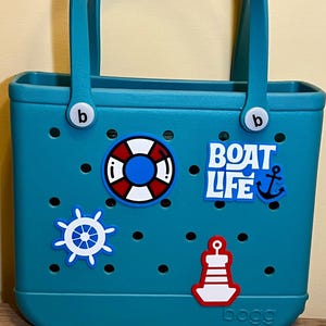 以下が含まれることがあります： 白と赤の航海をテーマにしたデカールが付いた、ティール色のトートバッグ。バッグにはハンドルがあり、側面に文字「b」があります。デカールには、救命浮き輪、船の舵、灯台、「BOAT LIFE」の文字と錨が含まれています。